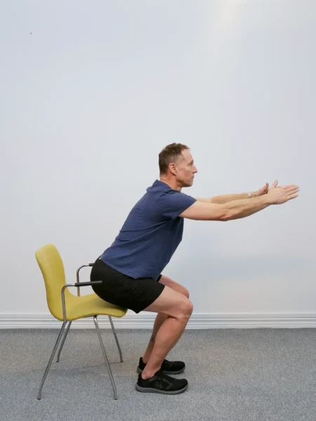 Person sänker sig kontrollerat i en knäböj med armarna rakt fram och höften bak mot stolen