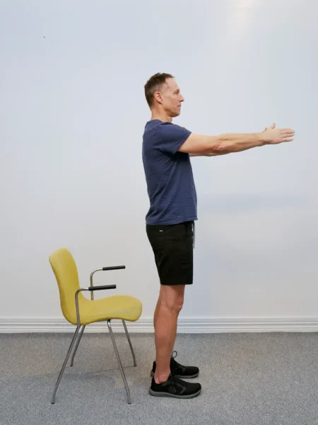 Person står upprätt med fötterna höftbrett, armarna utsträckta rakt fram och en stol placerad bakom kroppen