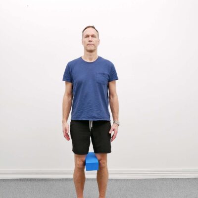 Person står upprätt med fötterna i höftbredd och en kudde placerad mellan knäna inför övningen standing knee squeezes.