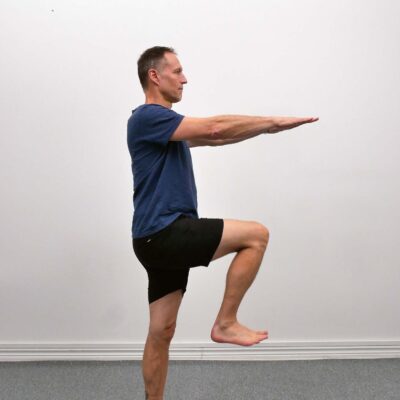 Person står upprätt med armarna sträckta framåt framför kroppen. Ett knä lyfts upp mot höftnivå medan överkroppen hålls upprätt och stabil.