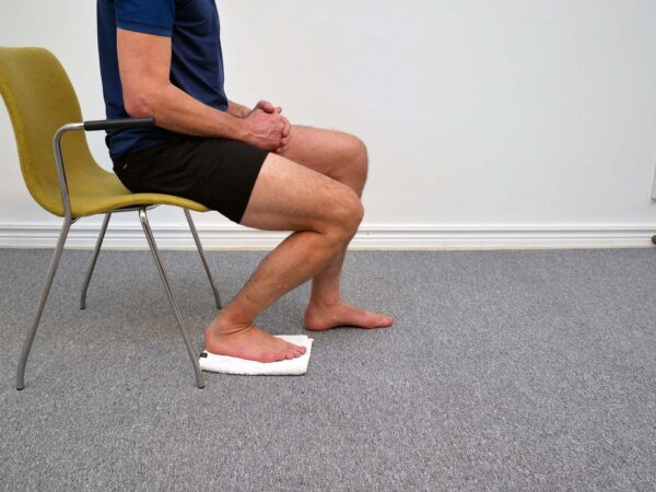 Person sitter på stol och har glidit foten framåt respektive bakåt för att visa rörelse i fotleden.