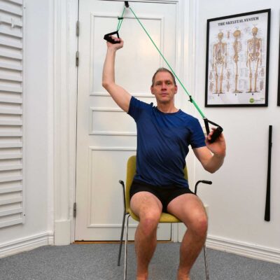 Slutposition: Person sitter på stol och har ena armen upplyft i axelabduktion med hjälp av slynga.