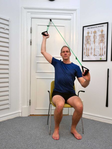 Slutposition: Person sitter på stol och har ena armen upplyft i axelabduktion med hjälp av slynga.