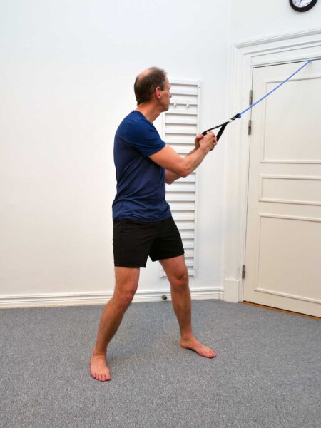 Person står brett isär och håller ett gummiband upp mot ena sidan av kroppen inför en diagonal bålrotation.