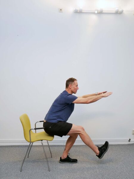 Personen sänker sig kontrollerat ned på ett ben med höften bakåt och knät böjt mot stolen bakom.