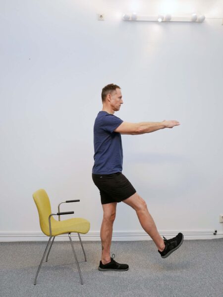 enbensknaboj-mot-stol-startposition: Person står på ett ben framför en stol med det andra benet sträckt framåt och armarna utsträckta.