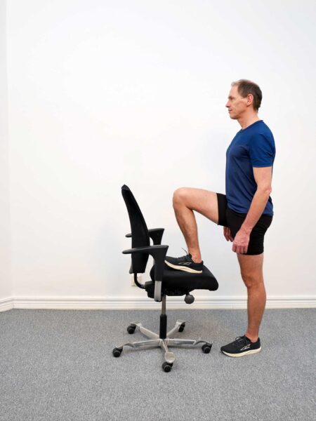 Person står bakom en stol med ena hälen placerad på sitsen, knät lätt böjt och blicken framåt.