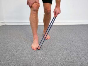 Person står med ena fotens stortå pressad ner mot golvet mot motståndet av ett gummiband. Slutposition för aktiv plantarflexion av stortåns flexor.