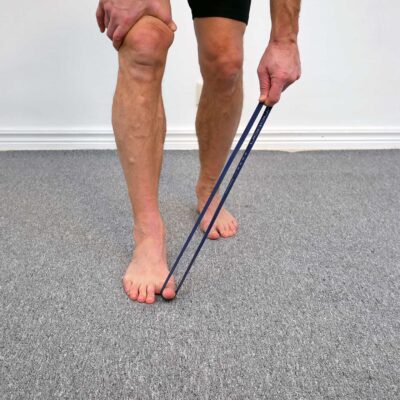 Person står med ena fotens stortå pressad ner mot golvet mot motståndet av ett gummiband. Slutposition för aktiv plantarflexion av stortåns flexor.
