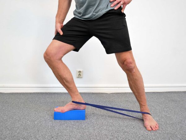 Person står med ena foten på ett yogablock, knät förs maximalt framåt över foten. Gummibandet runt talus drar språngbenet posteriort och underlättar djupare dorsalflexion i ankelleden.