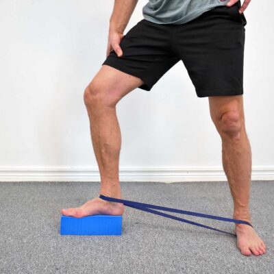 Person står med ena foten på ett yogablock. Ett gummiband är fäst runt språngbenet (talus) precis ovanför ankeln och sträcker bakåt för att dra talus posteriort vid dorsalflexion.