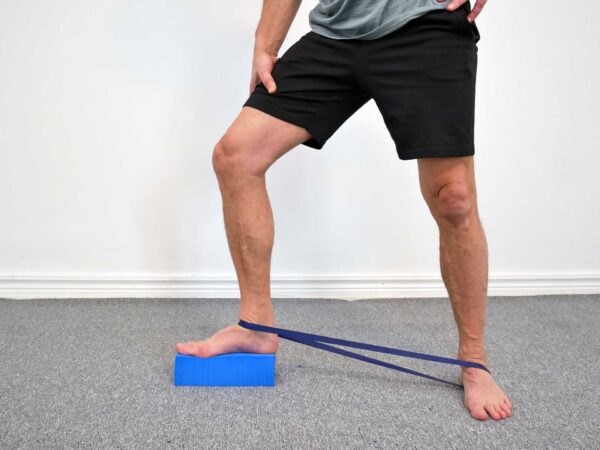 Person står med ena foten på ett yogablock. Ett gummiband är fäst runt språngbenet (talus) precis ovanför ankeln och sträcker bakåt för att dra talus posteriort vid dorsalflexion.