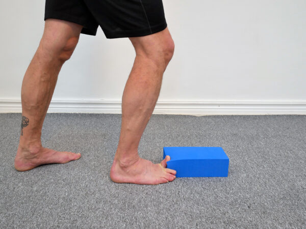 Person står med stortån upp på ett blått yogablock, hälen mot golv. Position för aktiv isometrisk nedåttryckning av stortån mot blocket för att stärka stortåns flexor.