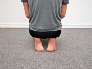 Person sittande på hälarna sett bakifrån. Båda fötter har tårna dorsalflexerade med framfoten mot golvet och hälarna upp mot sittbenen.