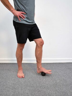 Person står med ena fotens fotvalv på en svart triggerboll, hälen mot golvet och tårna fria framför bollen - egenmassage av fotvalvets bindväv.