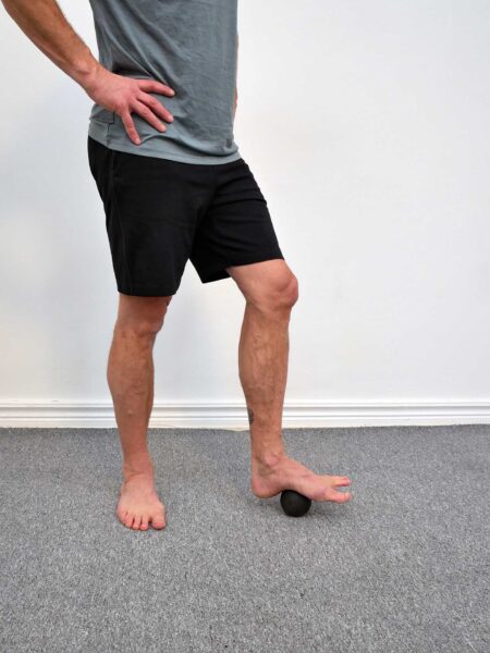 Person står med ena fotens fotvalv på en svart triggerboll, hälen mot golvet och tårna fria framför bollen - egenmassage av fotvalvets bindväv.