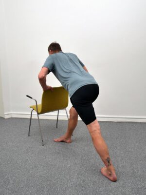 Djupare vadstretch i löpar-stretch-position med händerna på stolryggen - bakre häl trycks i golvet, fram-knä böjt