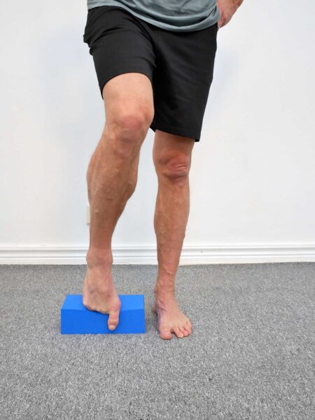 Person står upprätt med ena foten på ett blått yogablock och andra foten platt på golvet vid sidan, tårna avslappnade i naturligt läge på blockets ovansida.